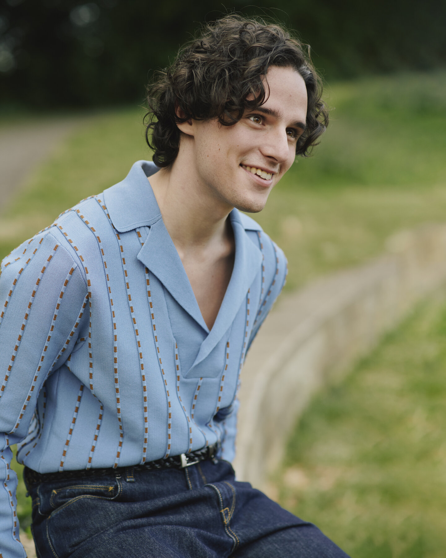 Harry Collett wears a blue prada jumper sitting on a wall with soft focus grassy background